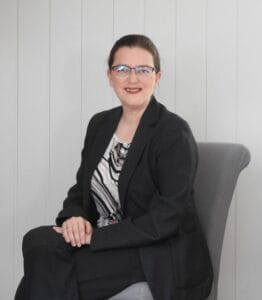 Portrait of Anastasia Olympiou-Hansen wearing glasses and a dark blazer, seated against a light background, based in Voss, Norway
