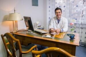 Man in a white medical coat seated at an office desk with a computer and documents at Dimitrios Pikos practice in Hürth