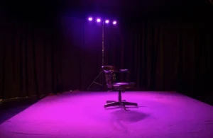 Empty theatre stage with black curtains, a single black chair and purple theatrical lighting, associated with Artion Theater in Munich