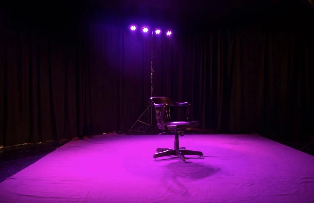 Empty theatre stage with black curtains, a single black chair and purple theatrical lighting, associated with Artion Theater in Munich